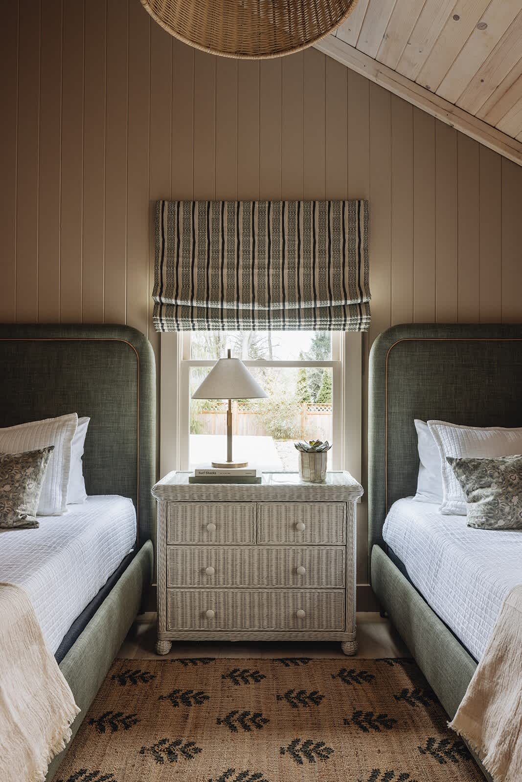 A close-up of a bedroom showing two twin beds with green upholstered headboards, separated by a white wicker nightstand with a lamp. The room features light brown paneling walls, a vaulted ceiling, and a patterned rug.