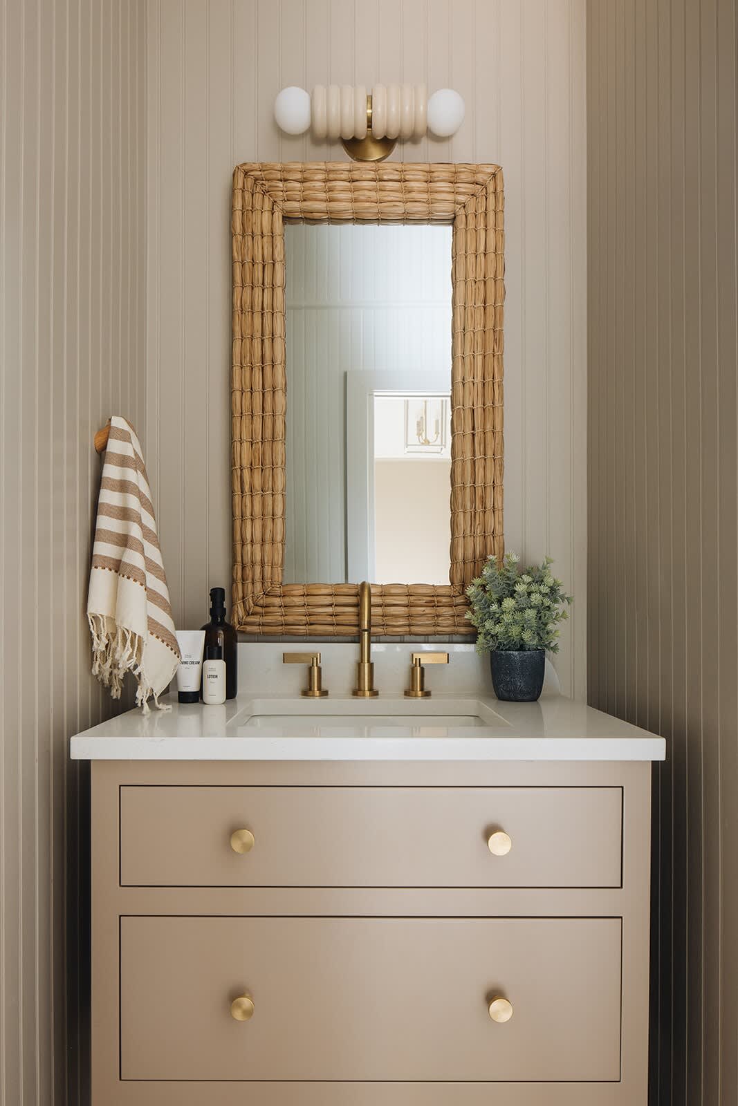 Stylish bathroom vanity area with light wood paneling, a beige vanity with gold knobs, a woven-framed mirror, and a modern wall sconce, featuring a gold faucet and white countertop.