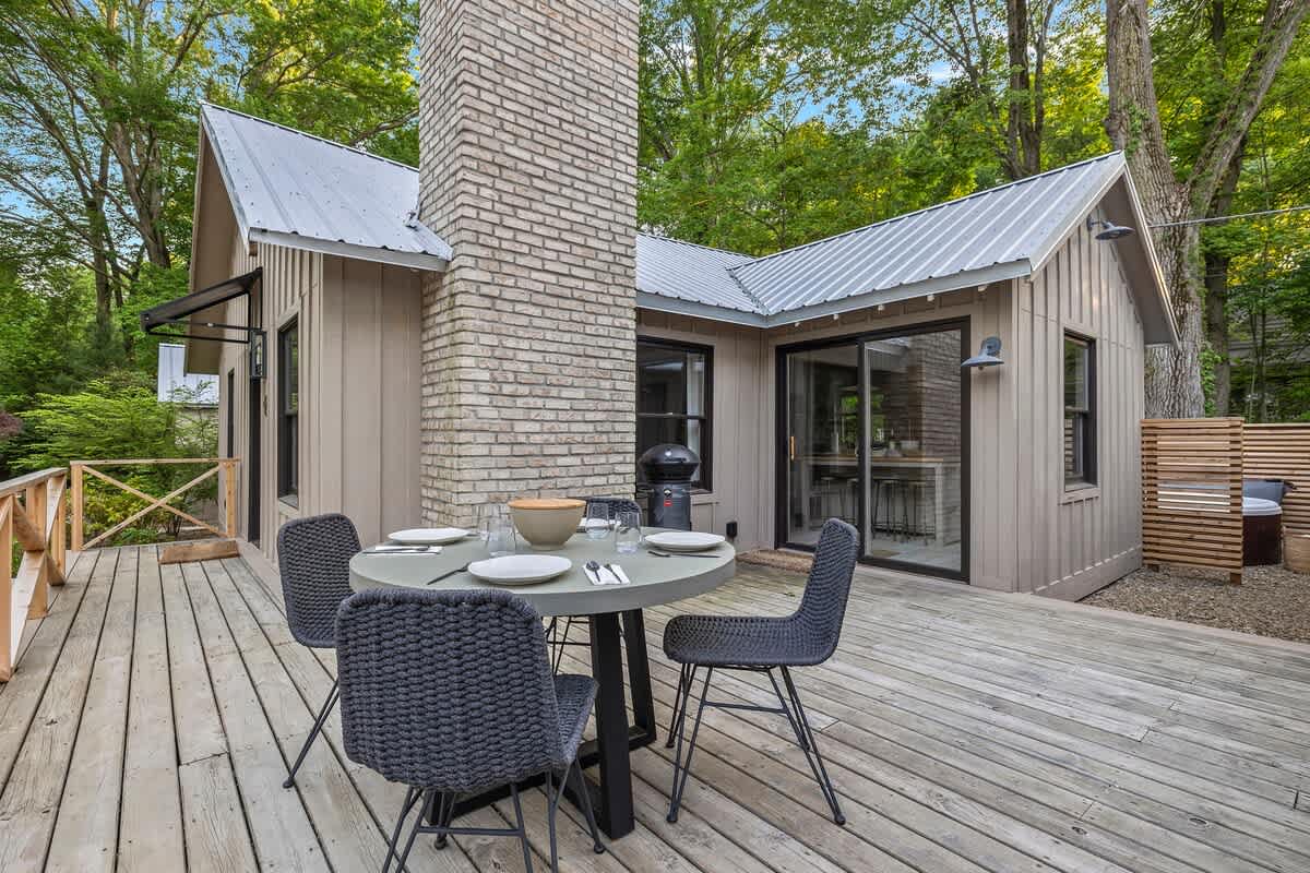 Expansive wooden deck with a round outdoor dining table, four chairs, and a BBQ grill, adjacent to the cottage with its metal roof and brick chimney, surrounded by greenery.
