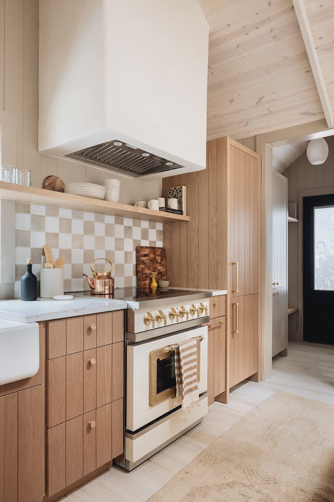 A view of the kitchen stove and range hood, featuring light wood cabinetry, a white and brass vintage-style range, a custom white range hood, and a checkered tile backsplash.