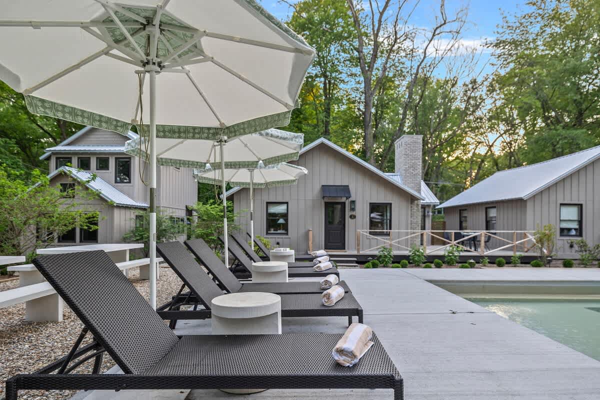 A poolside area with several dark lounge chairs, each with a rolled towel and a small side table, shaded by large white umbrellas, with modern light-colored cottages visible in the background.