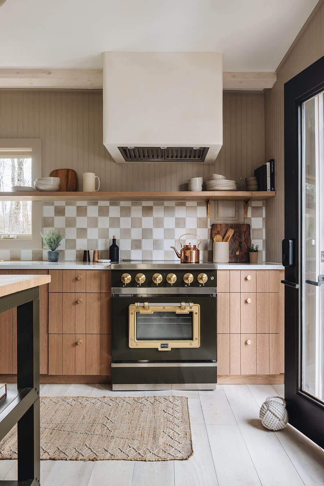 Detailed view of the kitchen featuring a dark green and brass professional-style range oven, a custom range hood, light wood cabinetry, and a white and grey checkered tile backsplash.