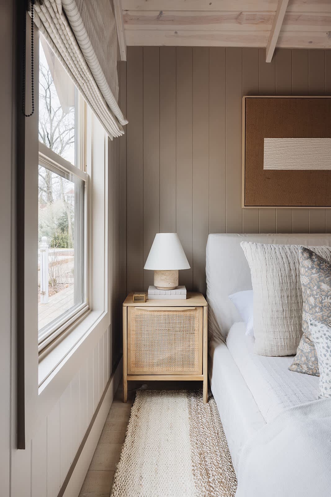 Close-up of a cane-front nightstand with a white ceramic lamp and stacked books, positioned next to a window with a striped Roman blind. The room features light beige vertical paneled walls and a textured rug.