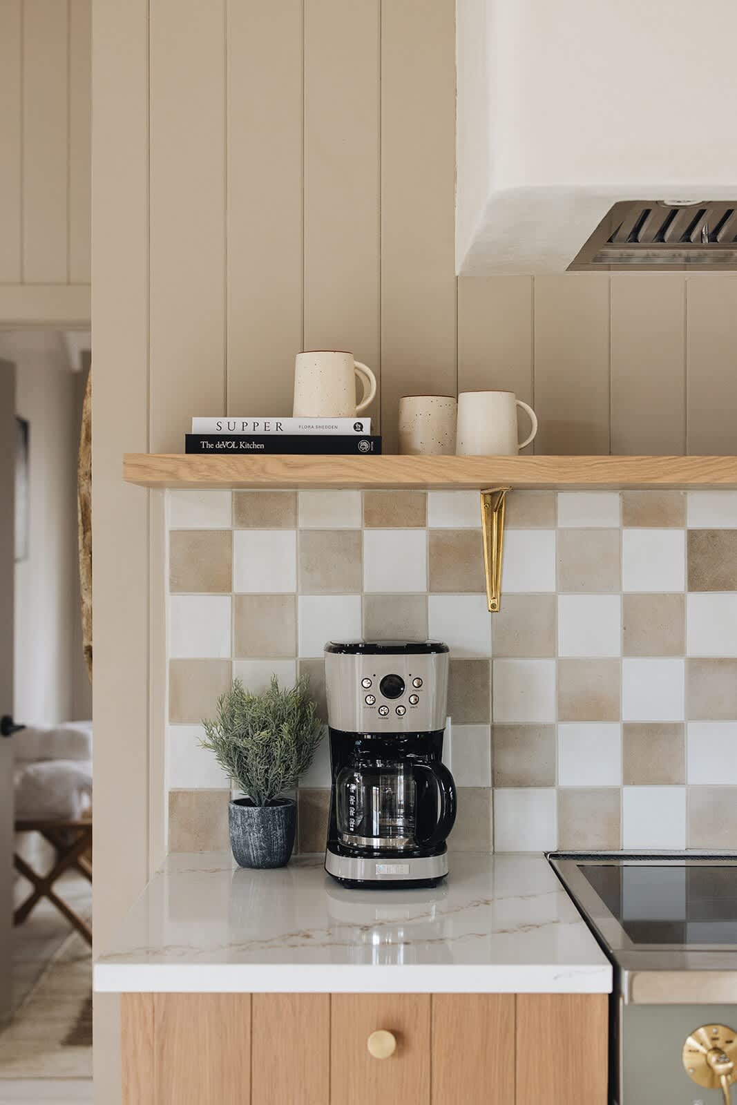 A section of the kitchen counter with a coffee maker, a small plant, and books on an open shelf, set against a checkered tile backsplash and light wood paneling.