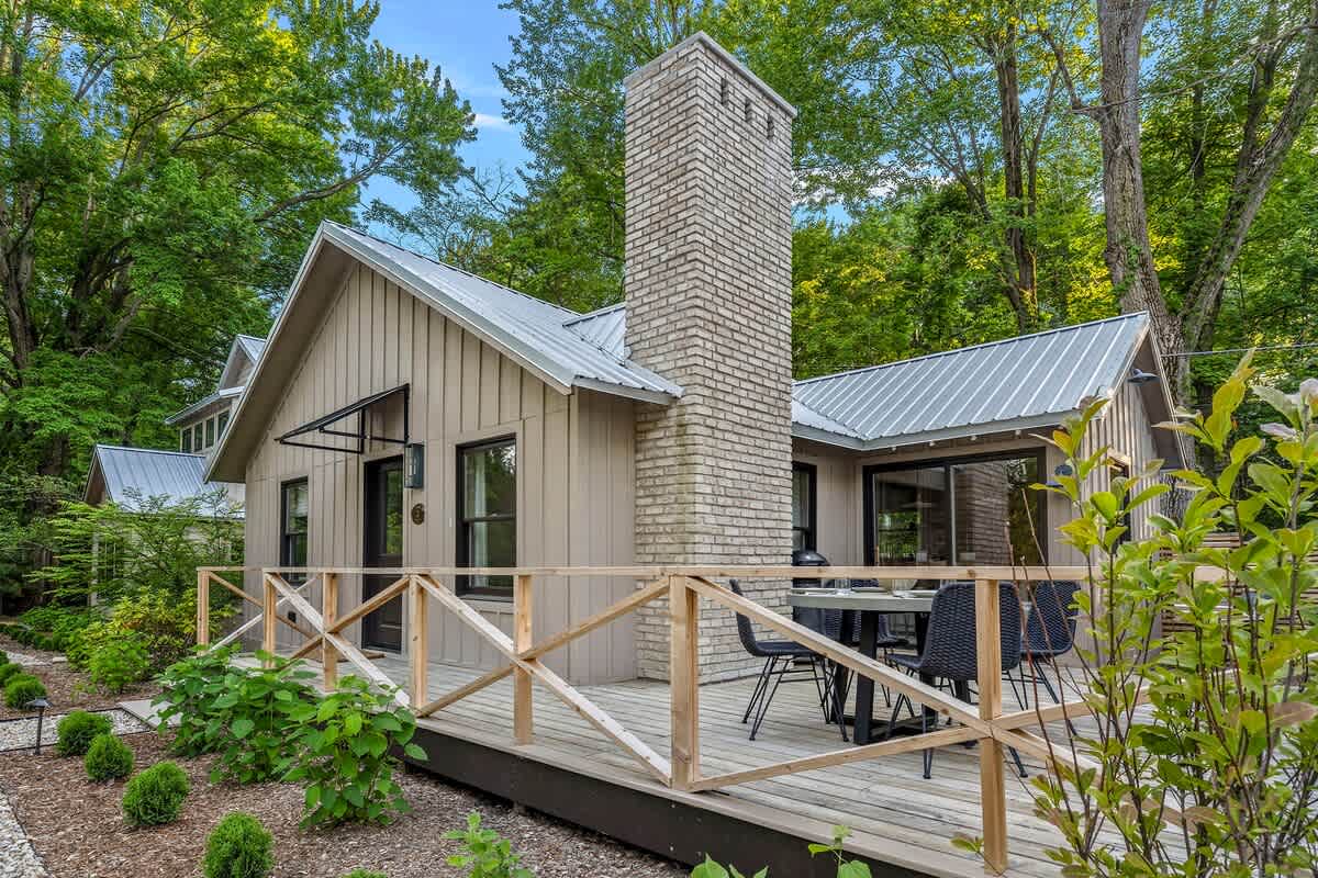 Side view of the cottage showcasing light beige board-and-batten siding, a metal roof, a prominent brick chimney, and a spacious wooden deck with a simple railing, surrounded by lush trees.