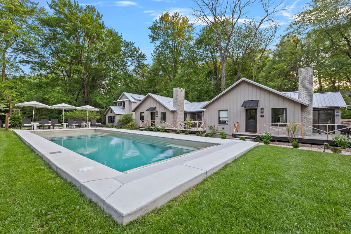 A serene outdoor swimming pool with clear blue water and steps, set within a concrete surround, flanked by a manicured green lawn and a row of modern light-colored cottages with dark roofs and brick chimneys, all under a bright sky.