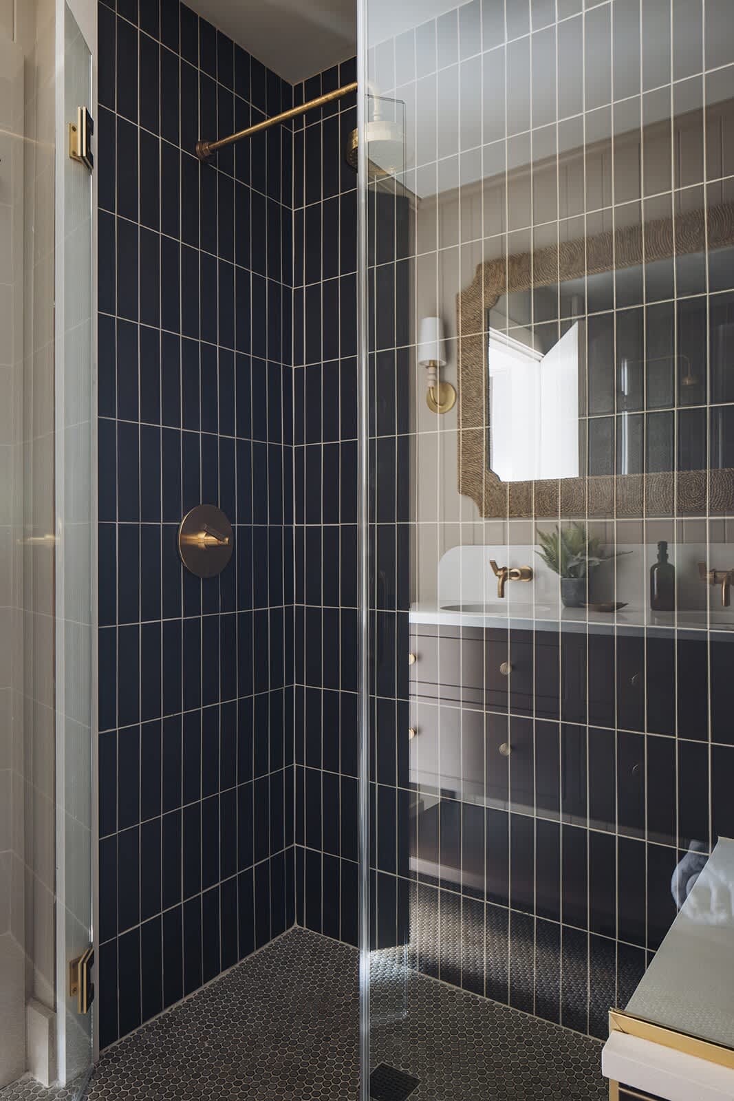 A modern shower stall with dark blue vertical subway tiles and a brass shower head and control. The floor is dark hexagonal tile, and a glass door separates it from the rest of the bathroom.