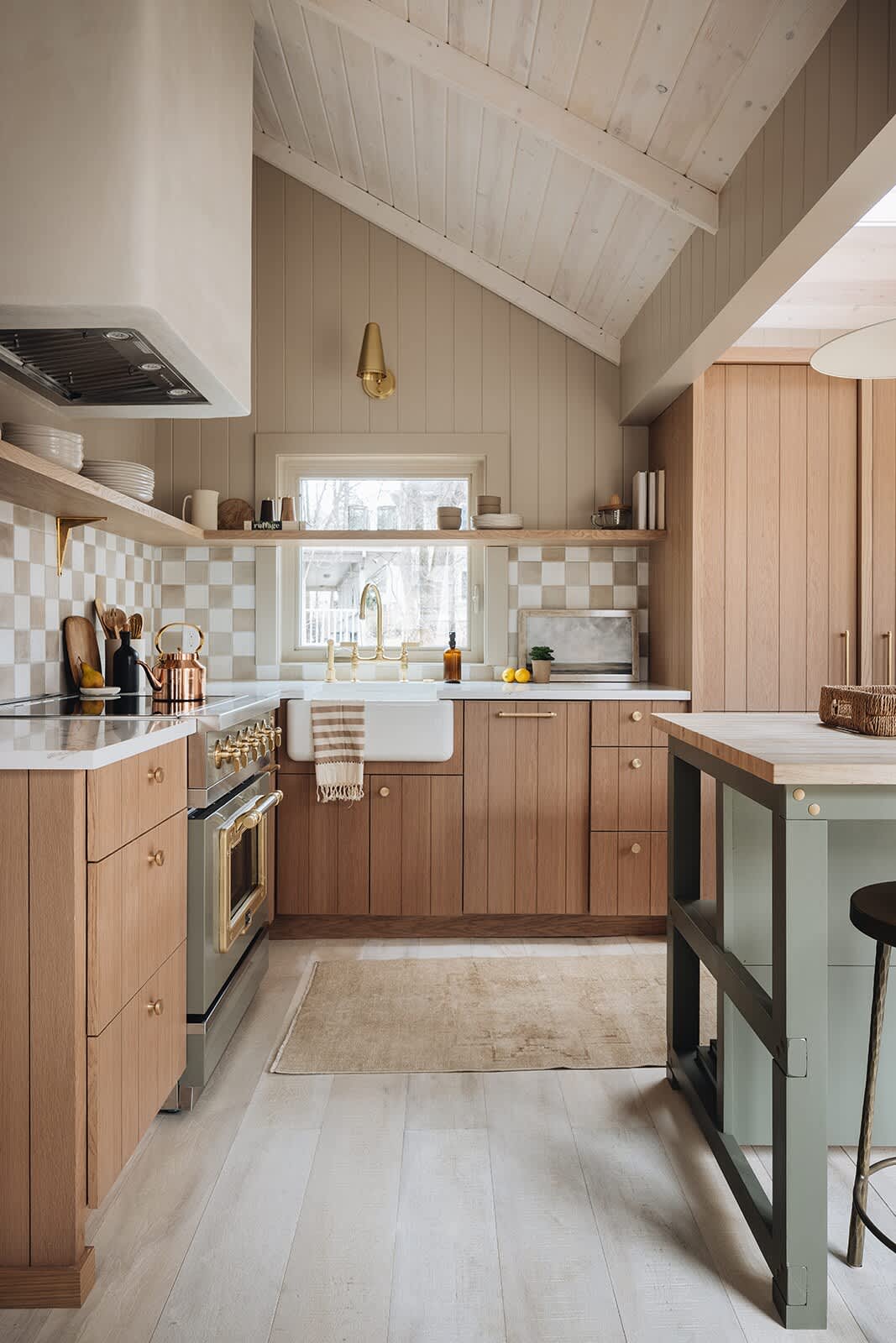 A cozy kitchen with light wood cabinets, a white farmhouse sink, and a checkered tile backsplash, featuring a sage green range and brass fixtures, under a vaulted wood ceiling.