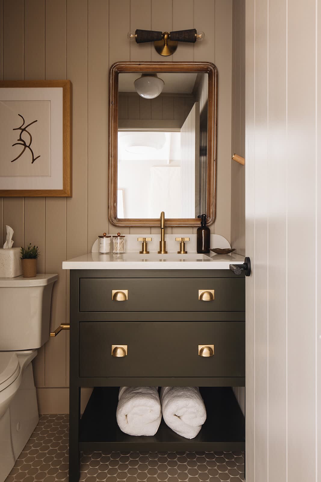 Close-up of a dark green vanity with brass drawer pulls and a white countertop, next to a white toilet, set against light beige vertical paneling and hexagonal floor tiles.