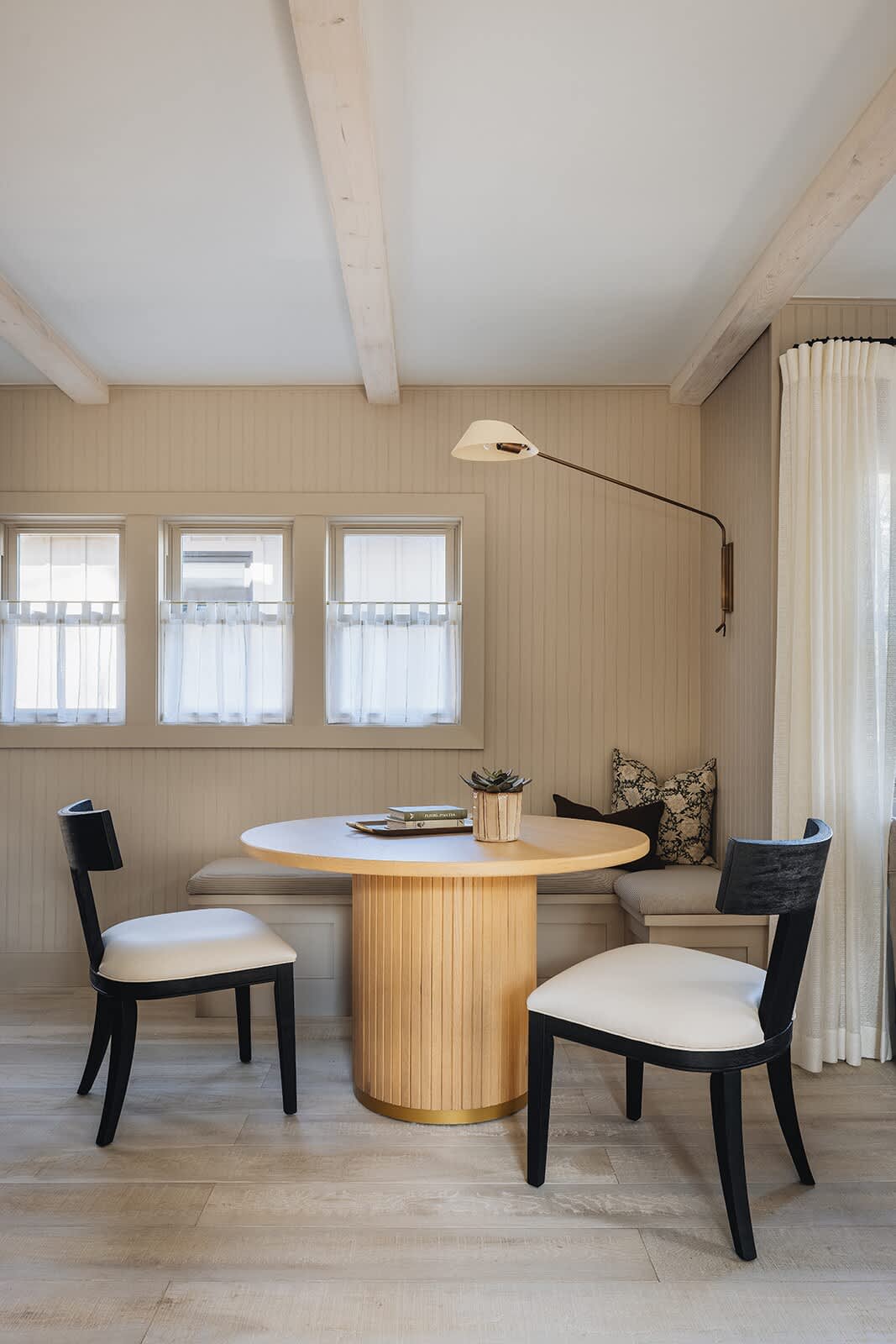 A bright dining nook with a round fluted wood table and two black and white chairs, complemented by a built-in bench seat. The room features light wood floors, exposed ceiling beams, and small windows with sheer curtains.