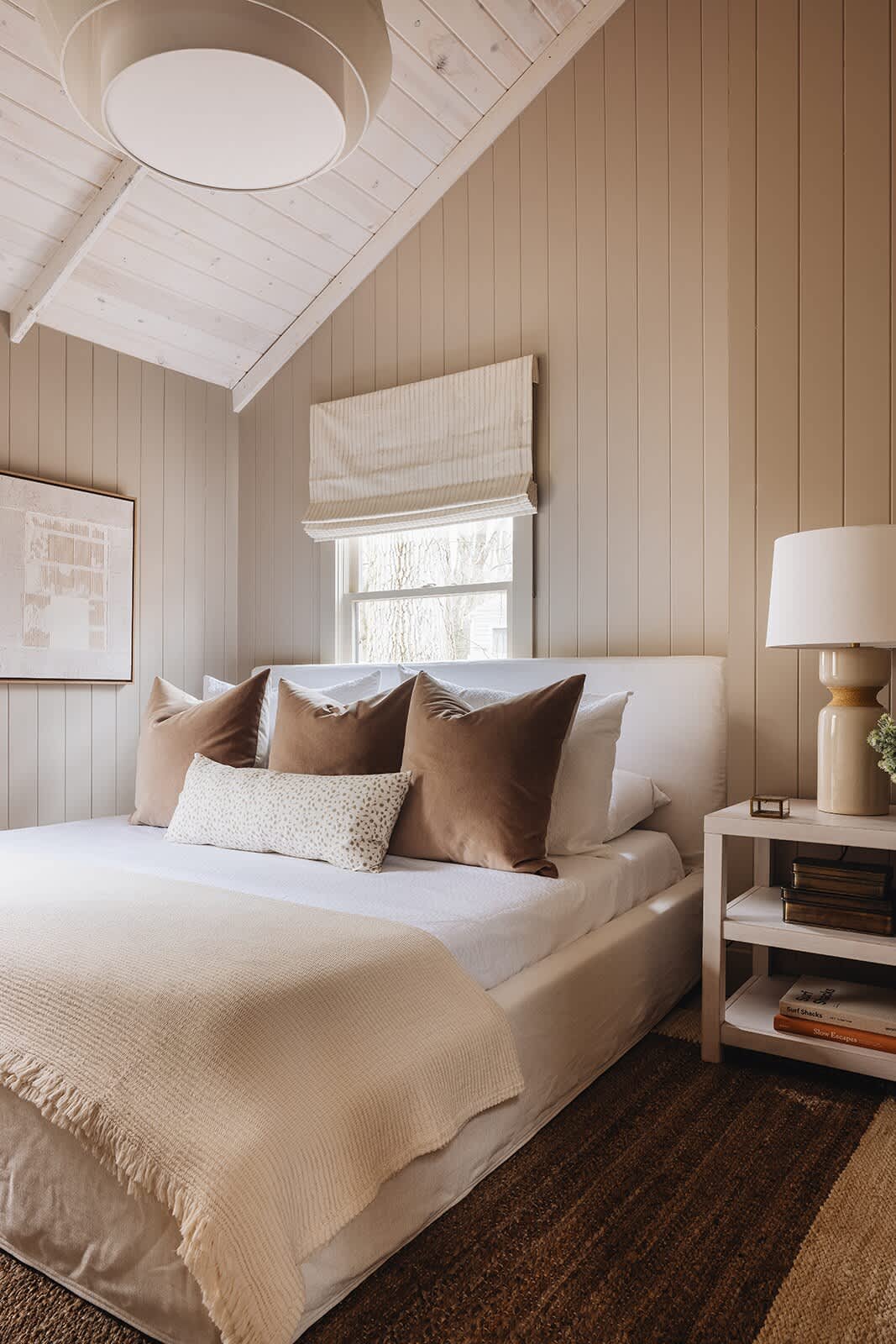 Cozy bedroom with a white upholstered bed, brown velvet pillows, a light wood nightstand with a lamp, and a sloped ceiling with white painted wood planks, all set against light beige vertical paneling.