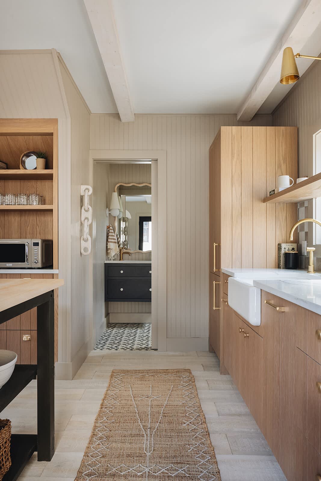 Transitional space with light wood paneling and flooring, featuring a built-in wooden shelving unit on the left and a view into a bathroom with a dark vanity and patterned floor tiles.