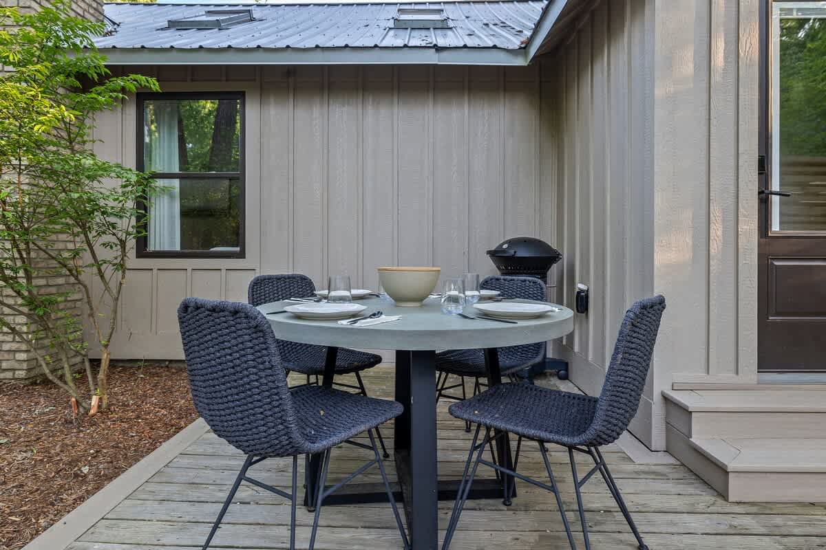 Outdoor dining area on a wooden deck next to a light beige cottage, featuring a round gray table set for four with dark woven chairs and a black charcoal grill.