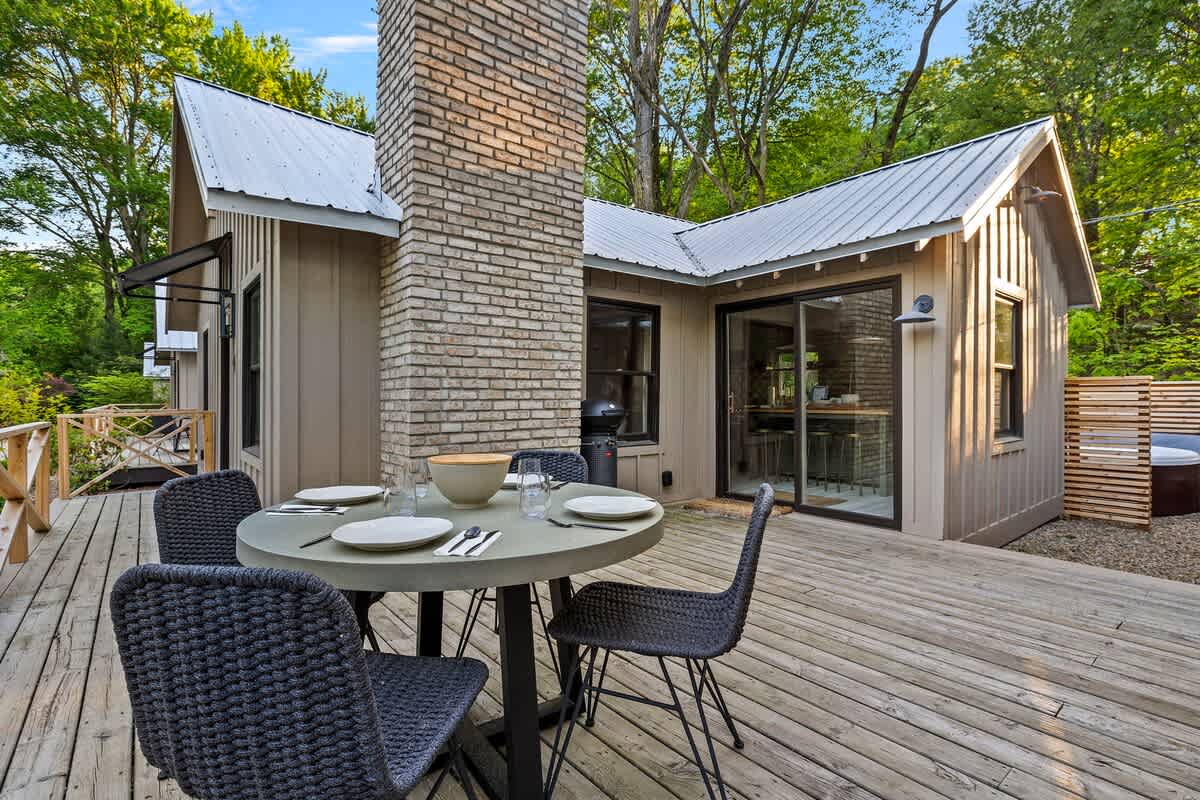 A spacious wooden deck with an outdoor dining set, positioned next to a light beige cottage featuring a large brick chimney and sliding glass doors. A grill is visible near the chimney, and a slatted wooden privacy screen hints at a hot tub area.