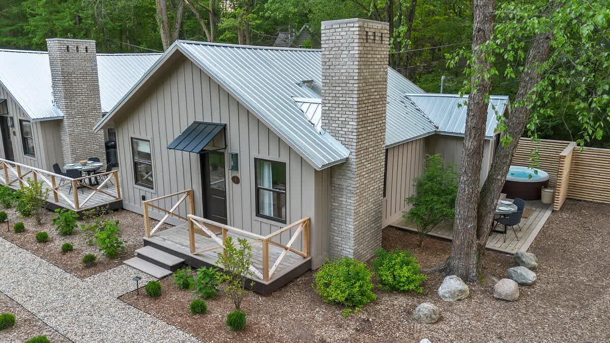 Aerial view of a modern cottage with light beige board-and-batten siding, a gray metal roof, and a prominent brick chimney. A gravel path leads to a wooden deck with a hot tub and outdoor dining area, surrounded by lush green trees.