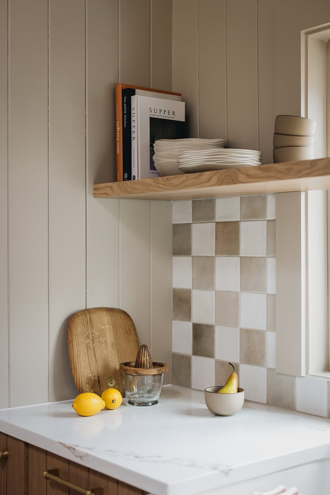 A detailed view of the kitchen counter with a white countertop, wooden cutting board, lemons, and a pear, positioned below an open wooden shelf and checkered tile backsplash.