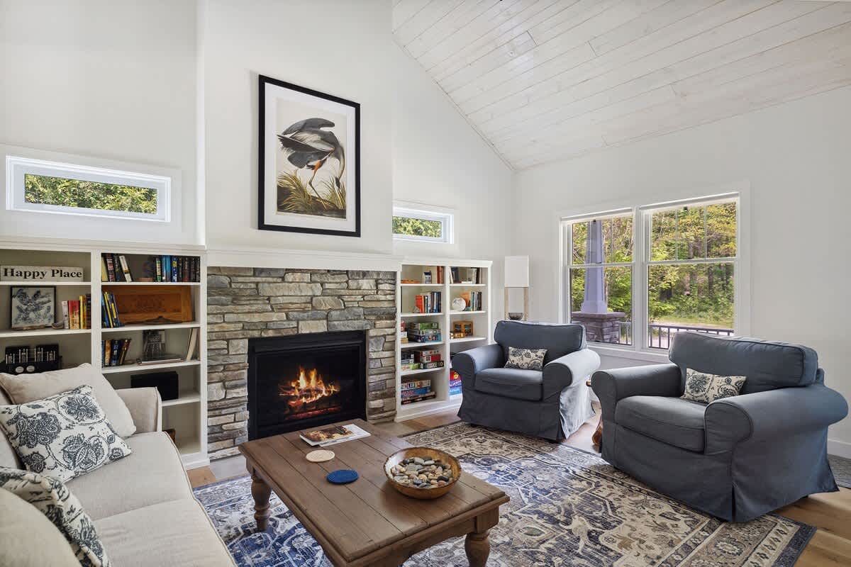 Inviting living room with a stone fireplace, two blue slipcovered armchairs, a light sofa, and a patterned rug, all under a high vaulted ceiling with white wood planks.
