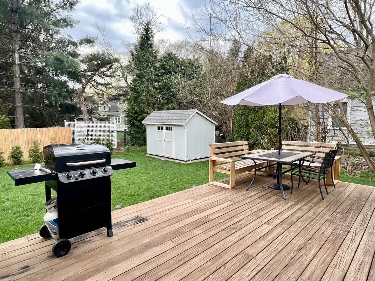 A spacious wooden deck featuring a black gas grill and an outdoor dining area with a white umbrella, wooden benches, and black chairs. A light blue shed is visible in the grassy backyard.