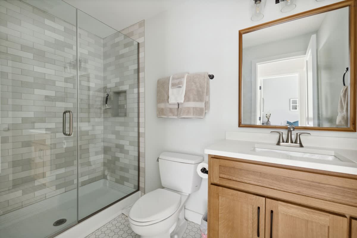 A modern bathroom featuring a glass-enclosed shower with light gray subway tile, a white toilet, and a light wood vanity with a white countertop and framed mirror. The floor has small hexagonal tiles.