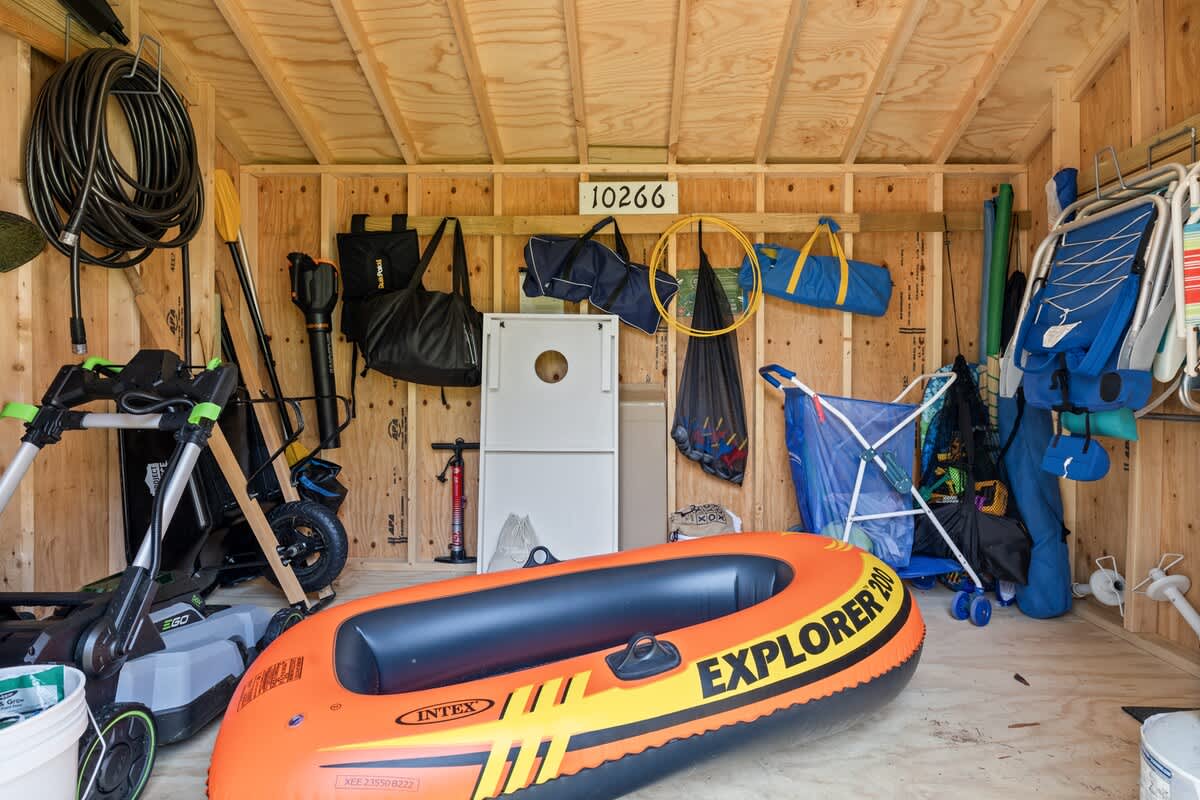 Wooden storage shed filled with various outdoor gear including an orange inflatable boat, a lawnmower, a cornhole set, and bags hanging on the walls.