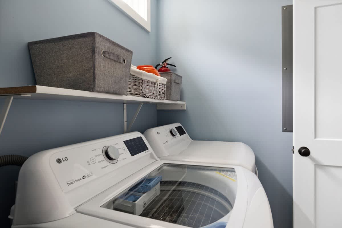 Bright laundry room featuring light blue walls, a white shelf holding storage baskets, and two white LG top-loading washing machines with clear lids, next to a white door.