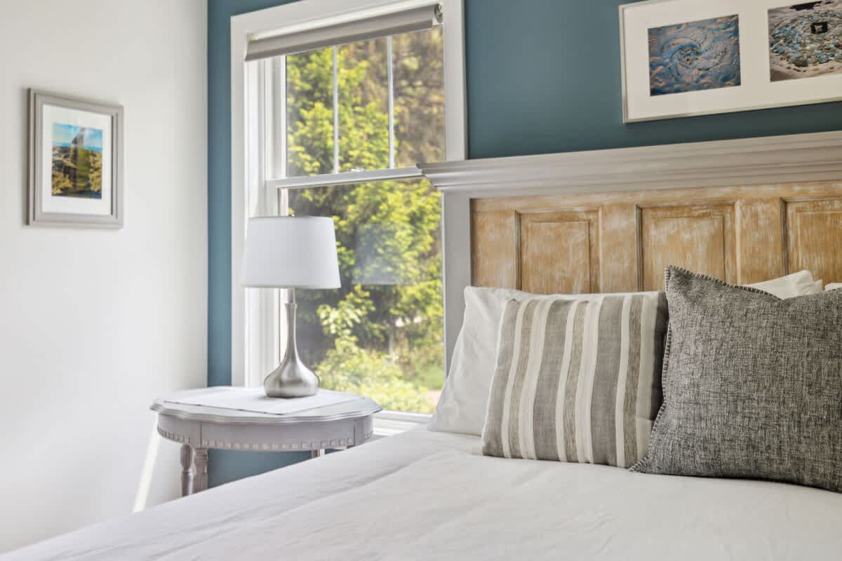 Close-up of a bedroom corner with a distressed wooden headboard, white bedding, striped and textured pillows, a round gray side table with a lamp, and a large window with a roller shade.