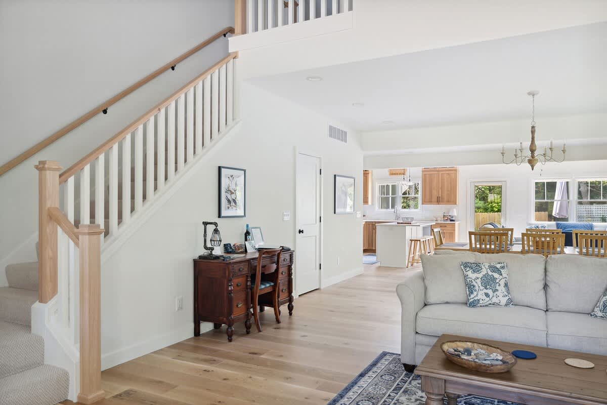 A bright foyer area featuring a wooden staircase with white balusters and a light wood handrail, leading upstairs, with a small antique-style wooden desk and chair below the stairs.