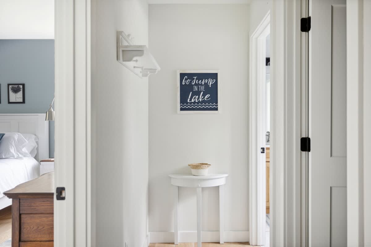 A bright white hallway with a small round white accent table and a "Go Jump in the Lake" framed art print. A glimpse of a bedroom with a blue accent wall and a bed is visible through an open doorway.