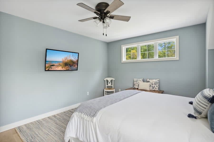 A bright bedroom with light blue walls, a large bed with white bedding and a gray throw, a wall-mounted television, a ceiling fan, and a window bench with pillows beneath a horizontal window.