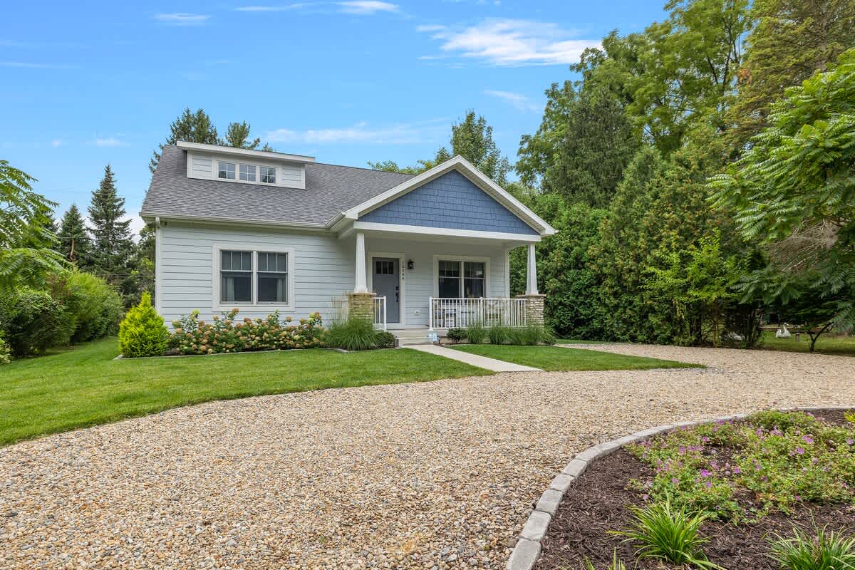 Daytime view of a charming light-sided house with a blue shingle gable, a welcoming front porch with white railings and stone pillars, and a curved gravel driveway leading to the entrance.