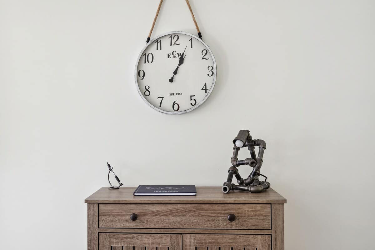 A decorative wall clock with rope hanger above a rustic wooden console table with drawers, featuring a unique industrial-style pipe lamp and a notebook, set against a plain white wall.