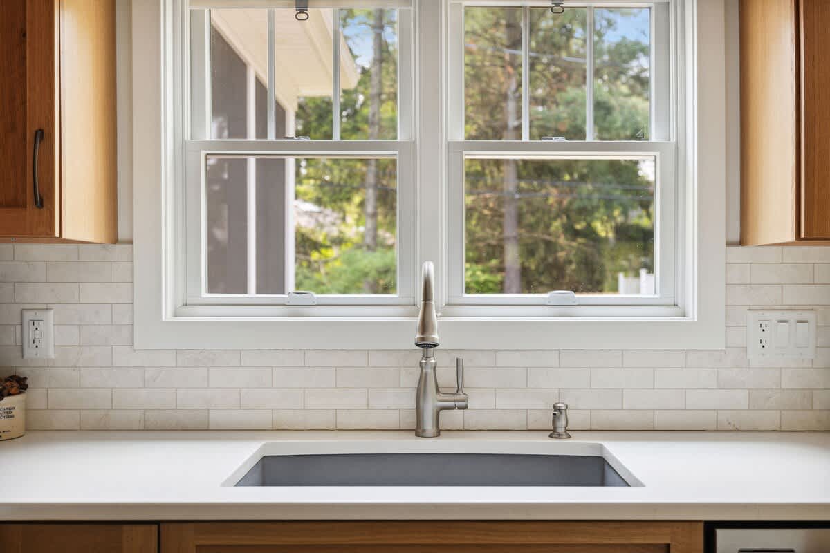 A close-up of the kitchen sink area with a stainless steel gooseneck faucet, a dark basin sink, white quartz countertops, and a white subway tile backsplash, beneath a large window overlooking greenery.