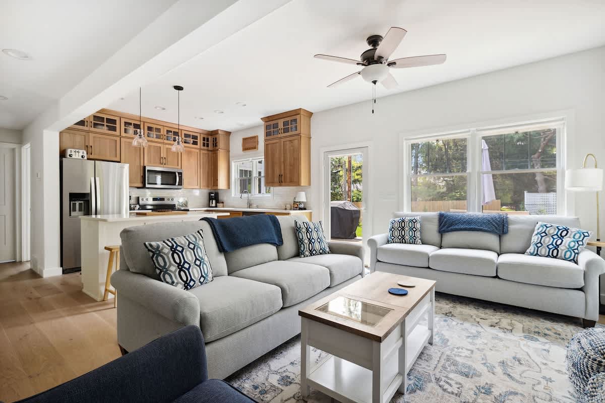 Open-concept living area featuring two light-colored sofas, a coffee table, and a view into a kitchen with wooden cabinets, stainless steel appliances, and a breakfast bar.