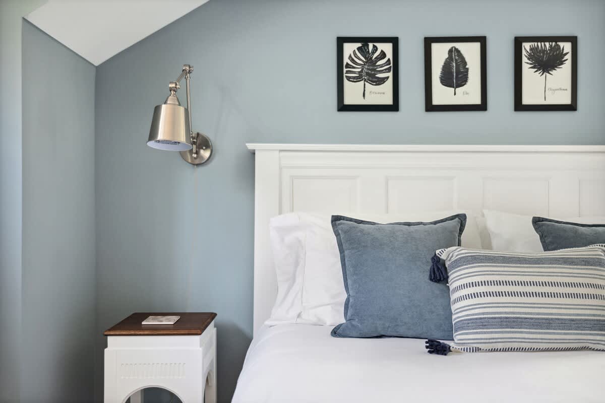 A cozy bedroom corner with a white paneled headboard, blue and striped throw pillows, a white nightstand, and a wall-mounted reading lamp, set against a light blue wall with framed botanical art.