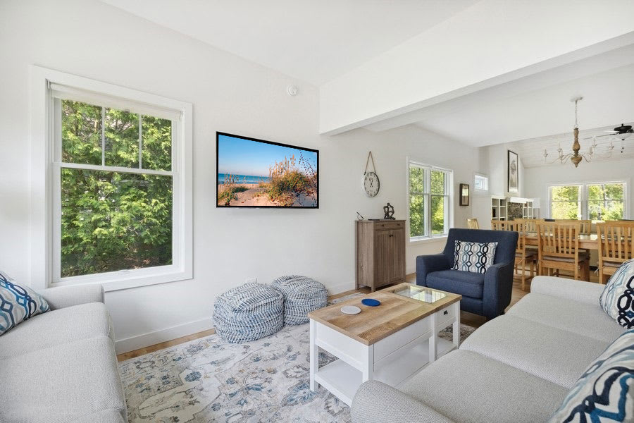 Bright living room with white walls, light wood floors, a large area rug, two light gray sofas, a blue armchair, and a wooden coffee table, featuring a wall-mounted TV and large windows overlooking greenery.