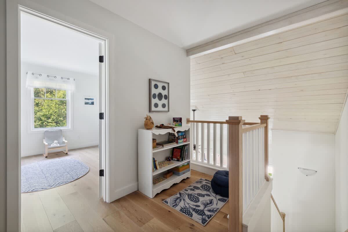 An upstairs landing with light hardwood floors, a white bookshelf filled with books and toys, a blue floor cushion, and a view into another room, with a sloped white wood-paneled ceiling.