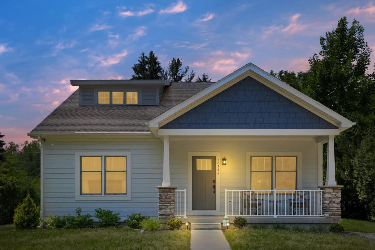 Evening view of the house with warm lights illuminating the front porch and windows, showcasing light siding, blue shingle accents, and a dark front door under a twilight sky.