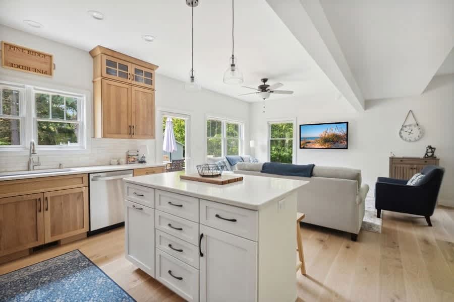 An open-concept kitchen and living area with a white kitchen island, light wood cabinets, white countertops, and light hardwood floors, leading into a bright living space with a sofa and wall-mounted television.