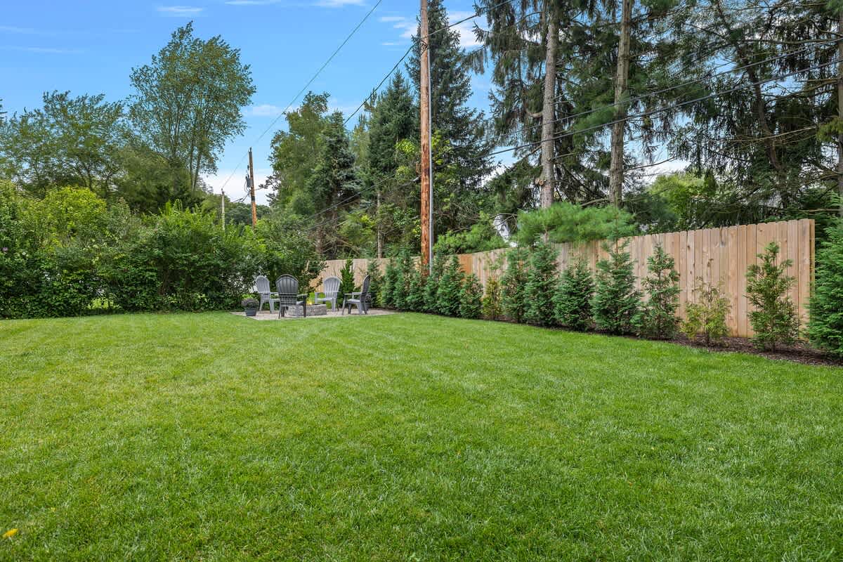 A wide view of a well-maintained green lawn in the backyard, with a wooden fence and tall trees in the background. The fire pit area with Adirondack chairs is visible in the distance.