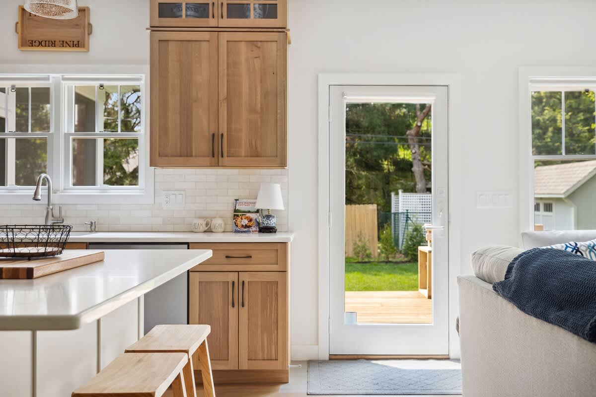 A bright kitchen area with light wood cabinetry, white countertops, and a white subway tile backsplash, featuring a kitchen island with stools and a glass-paned door leading to an outdoor deck.