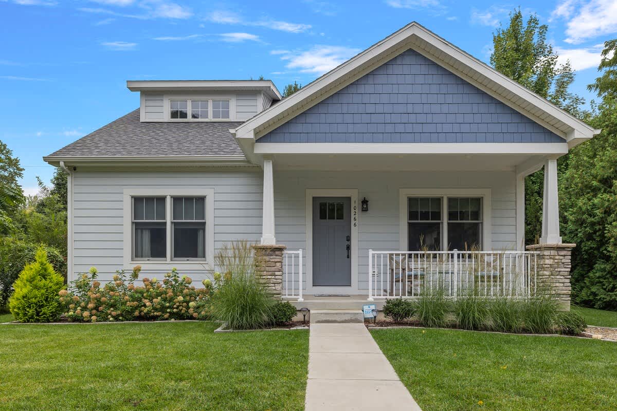 Charming light blue house with white trim, a covered front porch supported by stone pillars, and a well-maintained green lawn with lush landscaping.