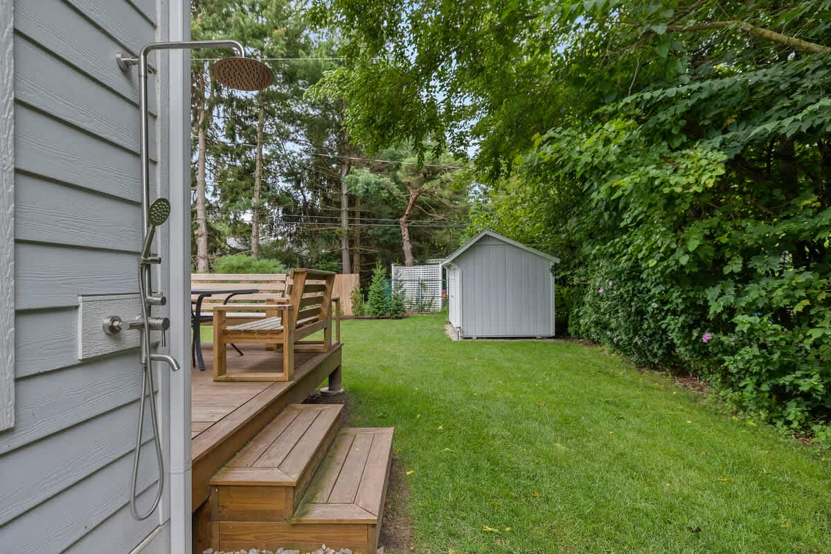 An outdoor shower with a rain showerhead mounted on the light blue siding of the house, adjacent to a wooden deck with steps leading to the grassy backyard. A shed is visible in the background.