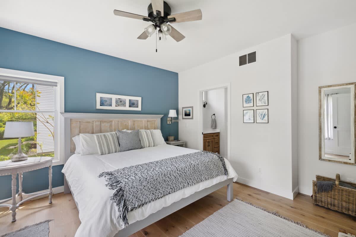Spacious bedroom with a king-sized bed, a blue accent wall behind the bed, light wood floors, a ceiling fan, and a large window, leading to an en-suite bathroom.