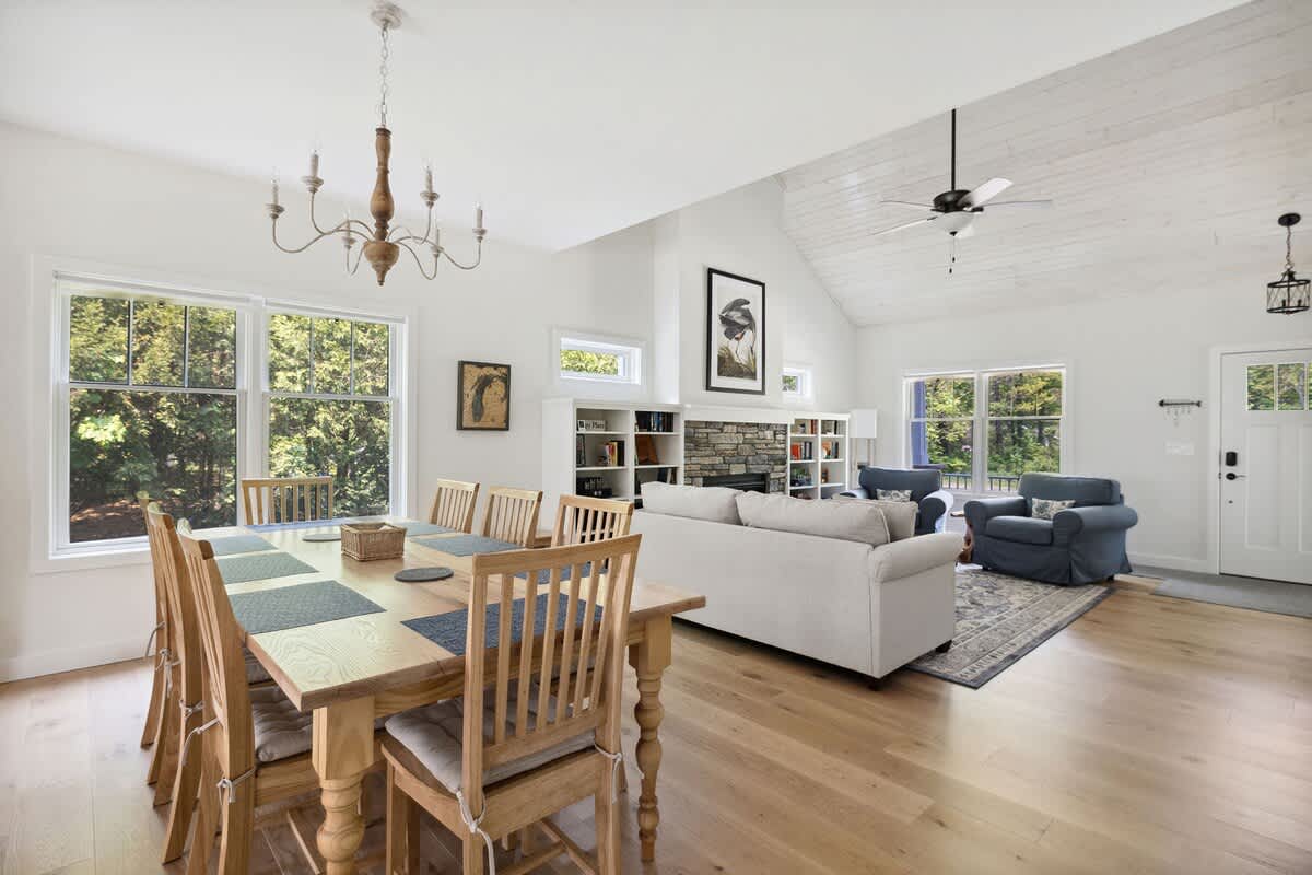 A spacious open-concept dining and living area with light hardwood floors, a large wooden dining table with matching chairs, and a living room beyond with a sofa, armchairs, and a stone fireplace.
