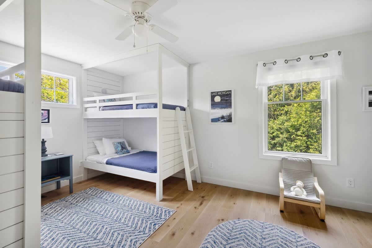 Kids' bedroom with white walls, light wood floors, a white bunk bed with blue bedding, a ceiling fan, and a large window, featuring a blue and white patterned rug.