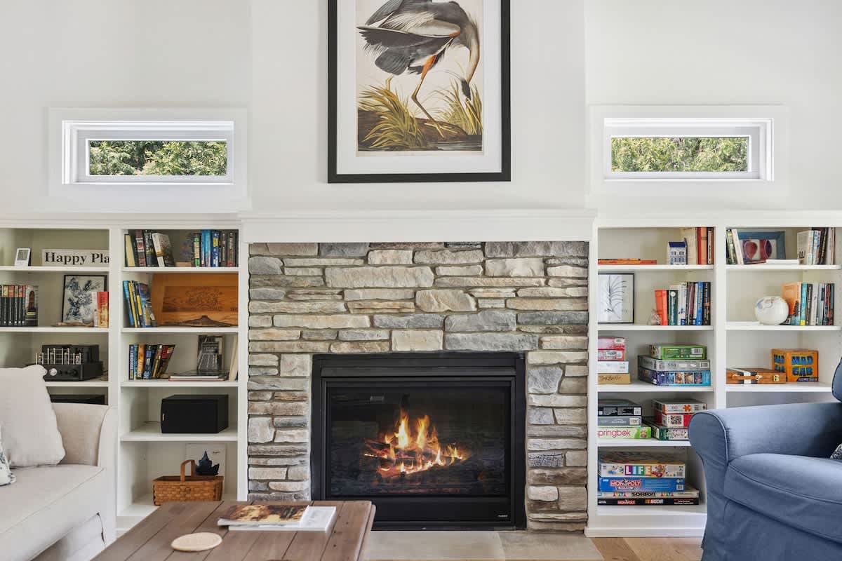 Cozy living room featuring a stone fireplace, flanked by white built-in bookshelves filled with books and board games, with light-colored seating and a high white ceiling.