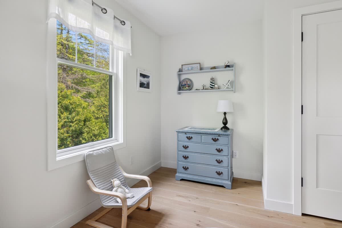 Corner of a kids' bedroom with a light blue dresser, a table lamp, a wall shelf with nautical decor, a gray rocking chair, and a large window overlooking greenery.