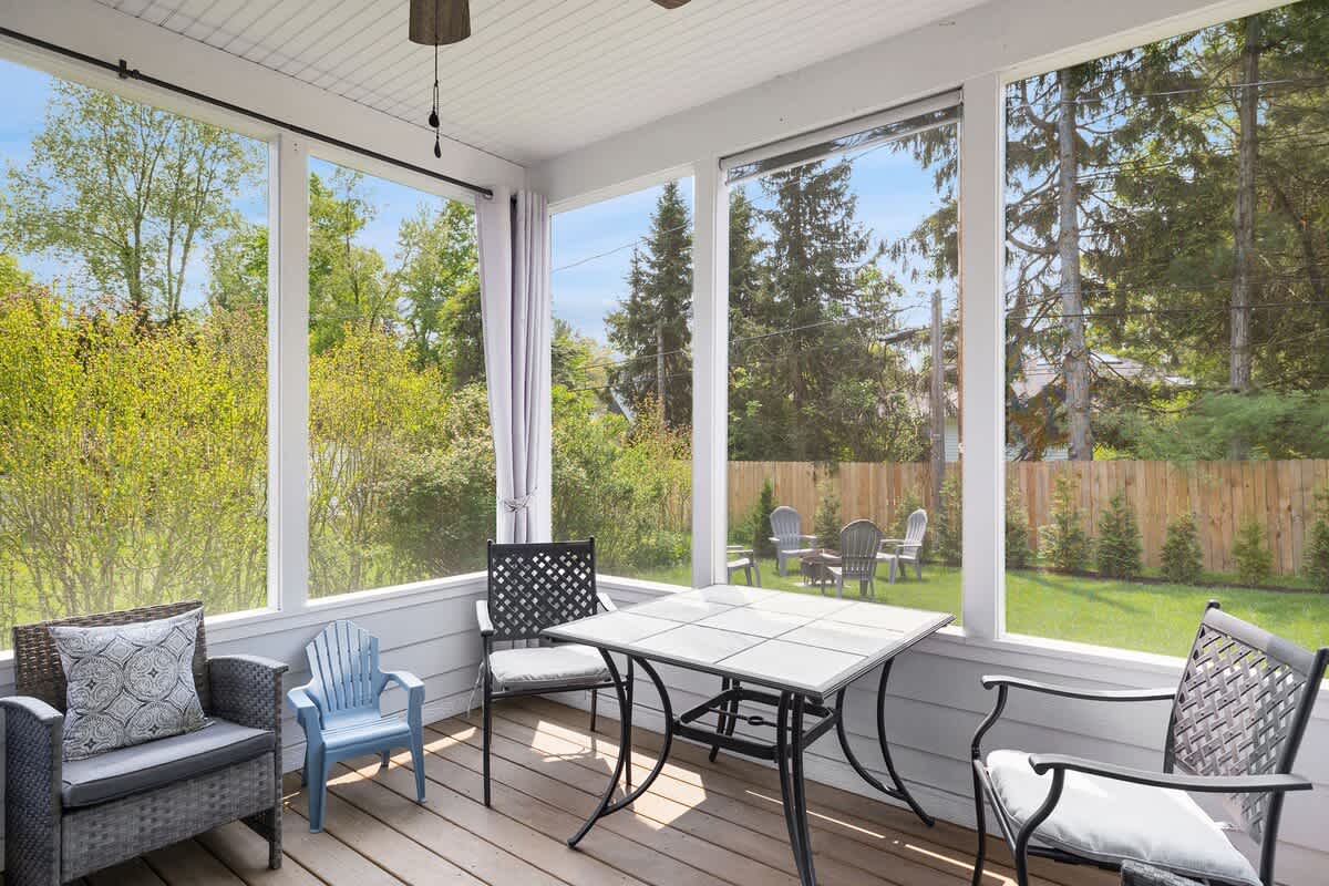 A cozy screened porch with a wooden floor, featuring a black metal dining table with chairs and a gray wicker armchair. Large windows offer views of a lush green backyard with a fire pit area.