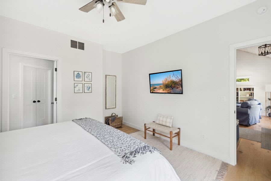 Bright bedroom with white walls, light wood floors, a king-sized bed with white bedding, a wall-mounted TV, a decorative bench, and a full-length mirror, with a doorway leading to another room.