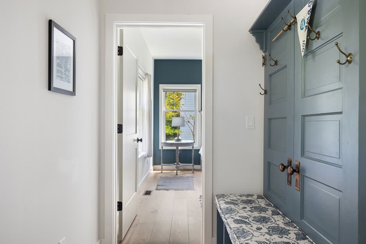 Bright hallway with light hardwood floors, a blue built-in coat rack and bench, and a doorway leading into a room with dark blue walls and a window.